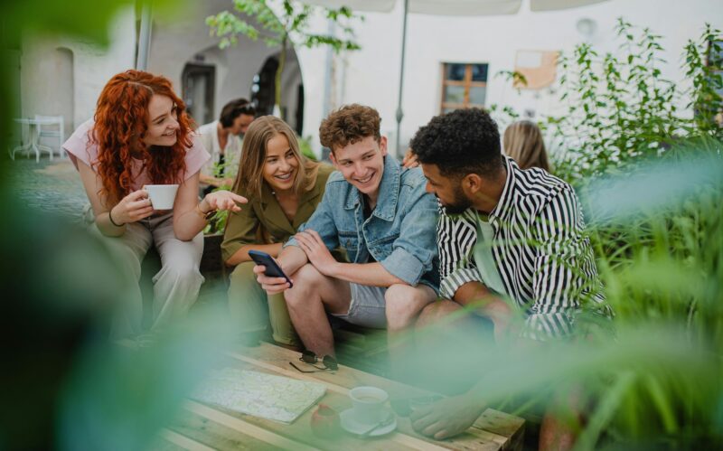 Group of happy young people with smartphone sitting in outdoors cafe on town trip, talking